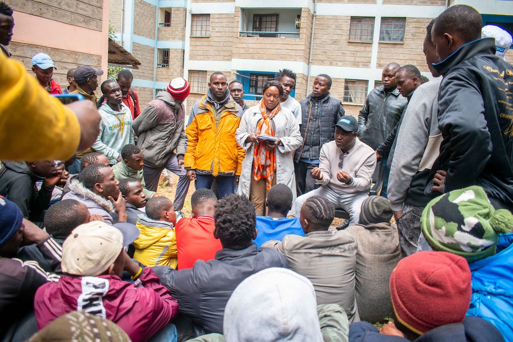 Shining Hope for Communities CEO Kennedy Odede speaks to community members who are sitting and standing around him. Their ages range from children to young people and older community members.