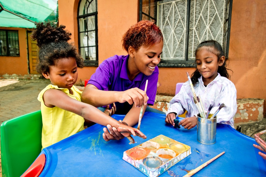 Early learning teacher painting with two young girl students at a blue table outdoors.