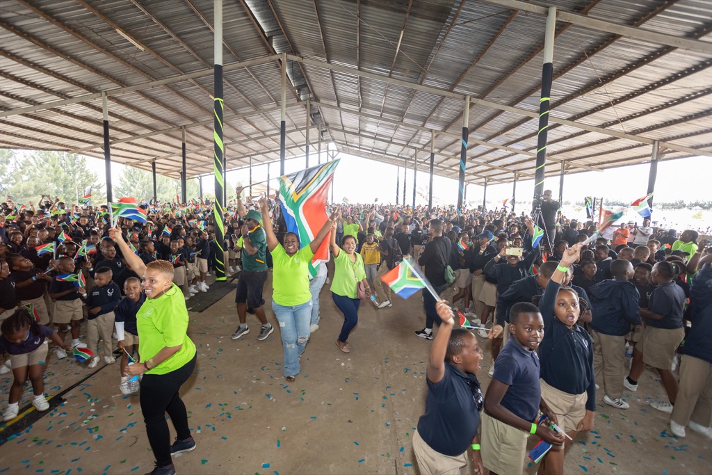 Spark Soweto School teachers are holding the South African flag and cheering with groups of students holding smaller South African flags. 