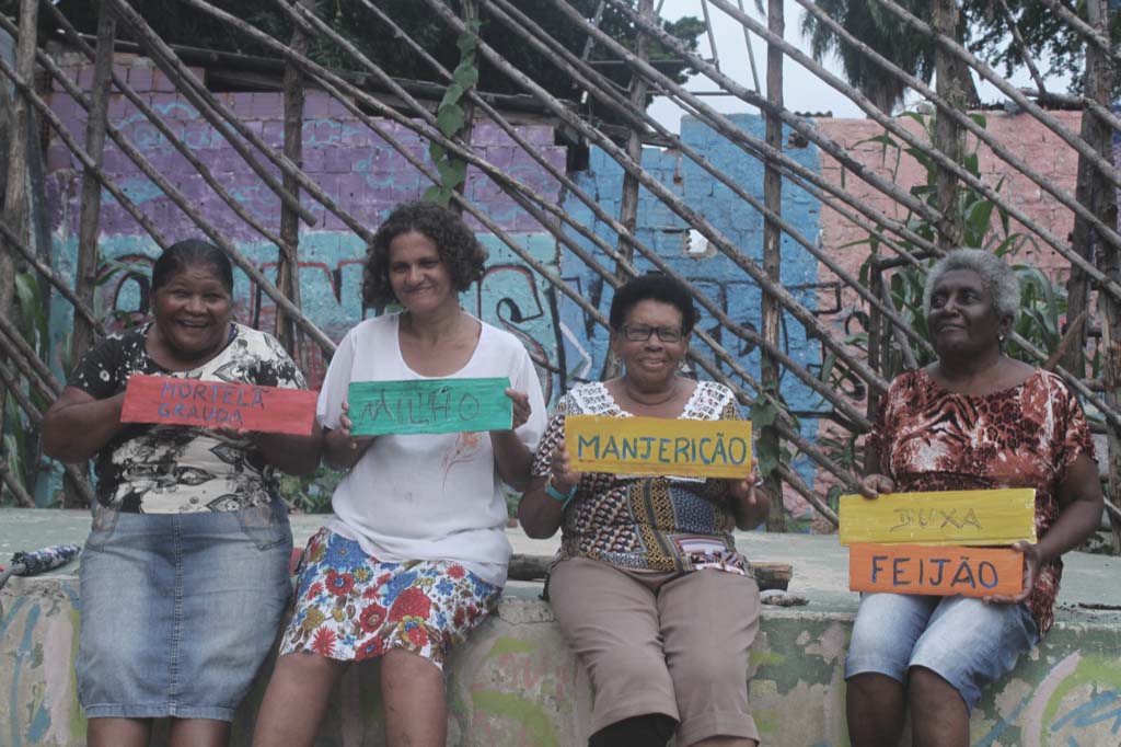 A group of four women sitting together. Each is holding a colorful sign that has the following words: "Hortela Grauda", "Milho", "Manjericao," and "Buxa Feijao".There is a colorful mural behind them.